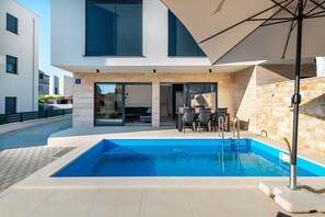 A modern outdoor pool area with a clear blue pool, a covered seating area with chairs and a table, and a stone wall. - Villa Sv. Josip (Vodice)