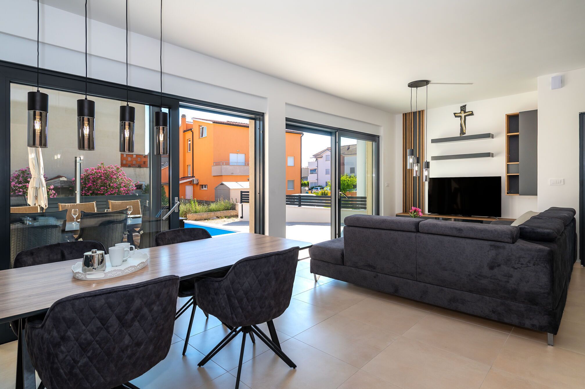 A modern living room with a grey sofa, a dining table with chairs, and a TV mounted on the wall.