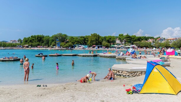 Ein Strand mit blauem Himmel, Menschen beim Schwimmen und Entspannen, und einem gelb-blauen Zelt im Sand.