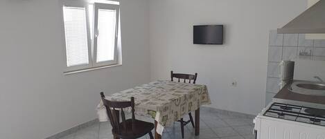 Une petite pièce propre, comprenant une table à manger, deux chaises, un téléviseur fixé au mur et un coin cuisine.