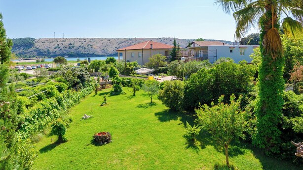 Μια οικιστική περιοχή με ένα μεγάλο σπίτι, πλούσια βλάστηση και θέα σε μια έκταση νερού και λόφους στο βάθος.