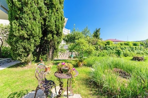 A garden with a table and chairs, a large tree, and a building in the background. - Apartments Ruzica M (Lopar)