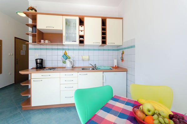A compact kitchen with white cabinets, a sink, and a table with a fruit basket. - Apartments Ruzica M (Lopar)