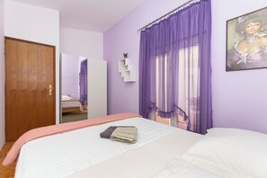 Ένα υπνοδωμάτιο με ένα κρεβάτι, μια ξύλινη πόρτα, μια λευκή ντουλάπα, μια μωβ κουρτίνα, ένα ράφι τοποθετημένο στον τοίχο με ένα φυτό και έναν πίνακα.