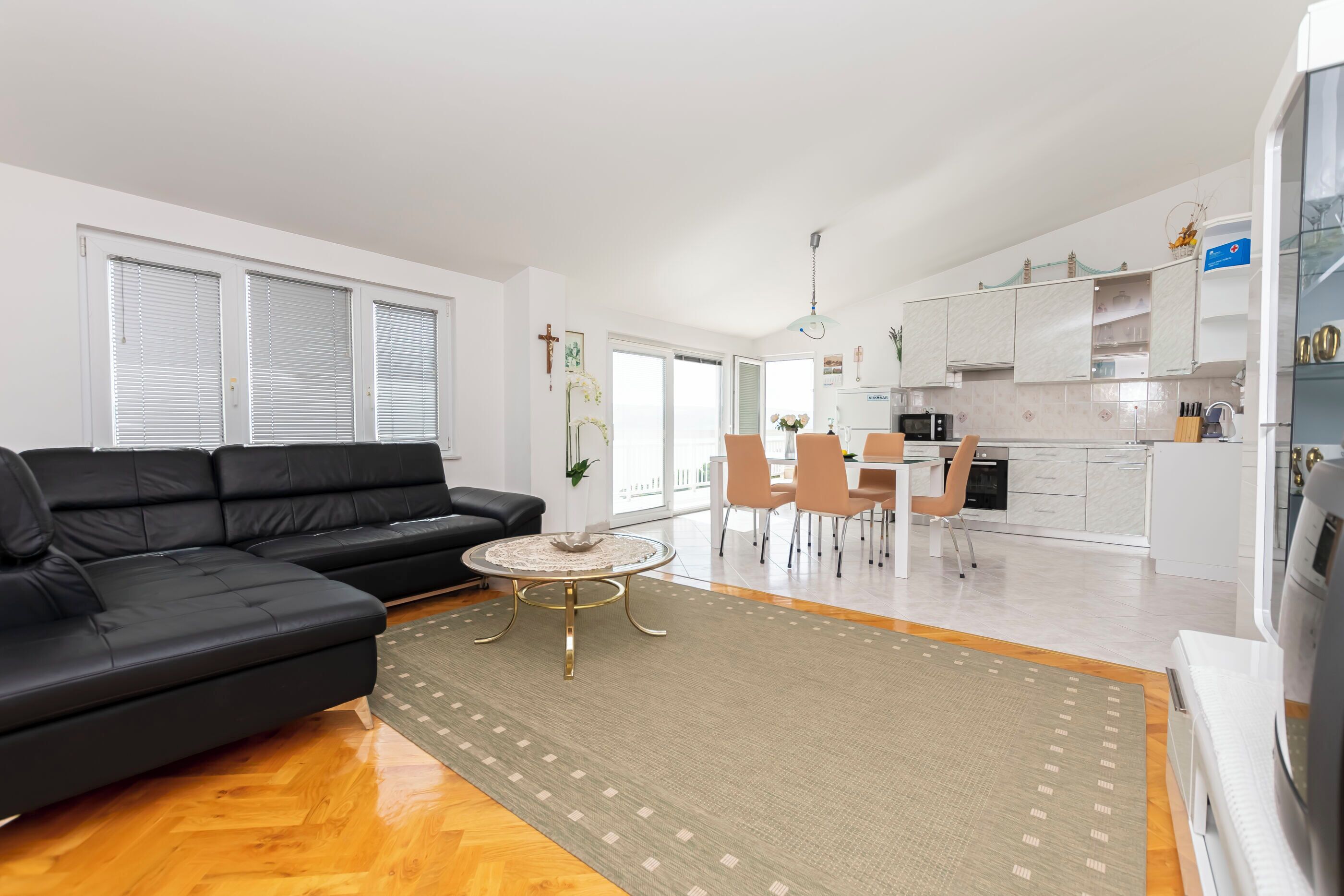 Una sala de estar moderna con un sofá negro, una mesa de centro redonda y un comedor con sillas y una mesa. Se ve la cocina al fondo, con armarios blancos y un microondas.