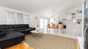 Una sala de estar moderna con un sofá negro, una mesa de centro redonda y un comedor con sillas y una mesa. Se ve la cocina al fondo, con armarios blancos y un microondas.