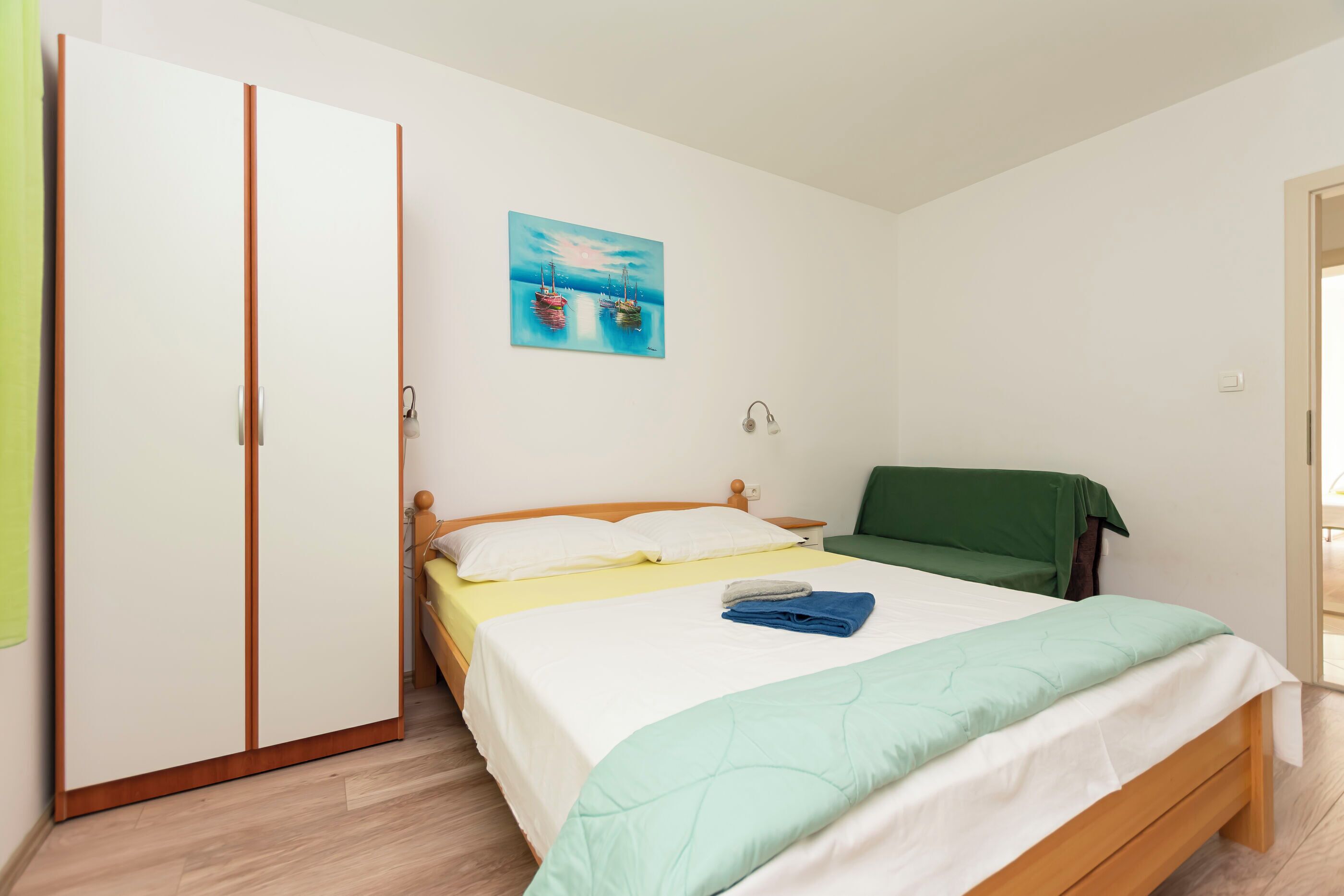 A bedroom with a bed, a green chair, a painting, and a wardrobe.