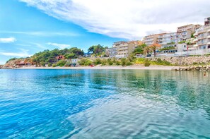 Μια παραθαλάσσια πόλη με κτίρια που βρίσκονται κατά μήκος της προκυμαίας, κρυστάλλινα, γαλάζια νερά και μια βραχώδη ακτογραμμή.