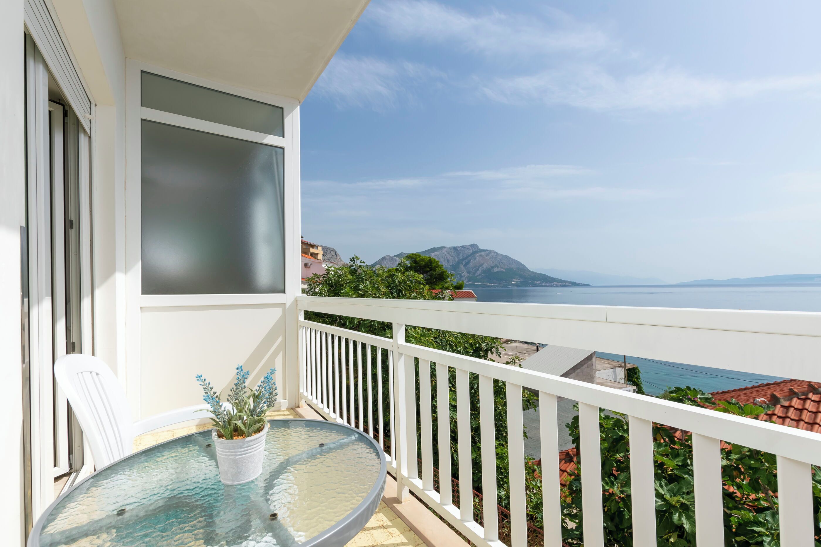 A balcony with a glass table and chairs, overlooking a coastal view with mountains and the sea.