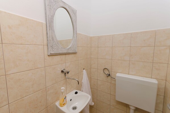 Une salle de bain avec des carreaux de couleur beige, un lavabo blanc doté d’un distributeur de savon, un miroir avec un cadre orné et des toilettes avec une cuvette en métal.