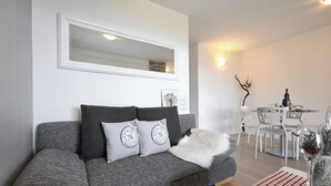 Una sala de estar moderna con un sofá gris, un plaid blanco suave y una zona de comedor con sillas blancas y una mesa.