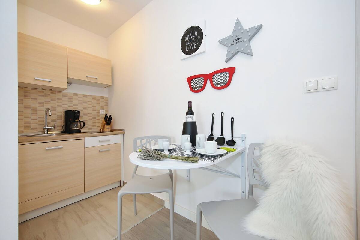 Une cuisine compacte avec des meubles en bois, un plan de travail blanc et un coin repas comprenant une table et des chaises blanches. Les murs sont décorés, notamment avec une étoile et une paire de lunettes de soleil, et il y a un fauteuil blanc moelleux.