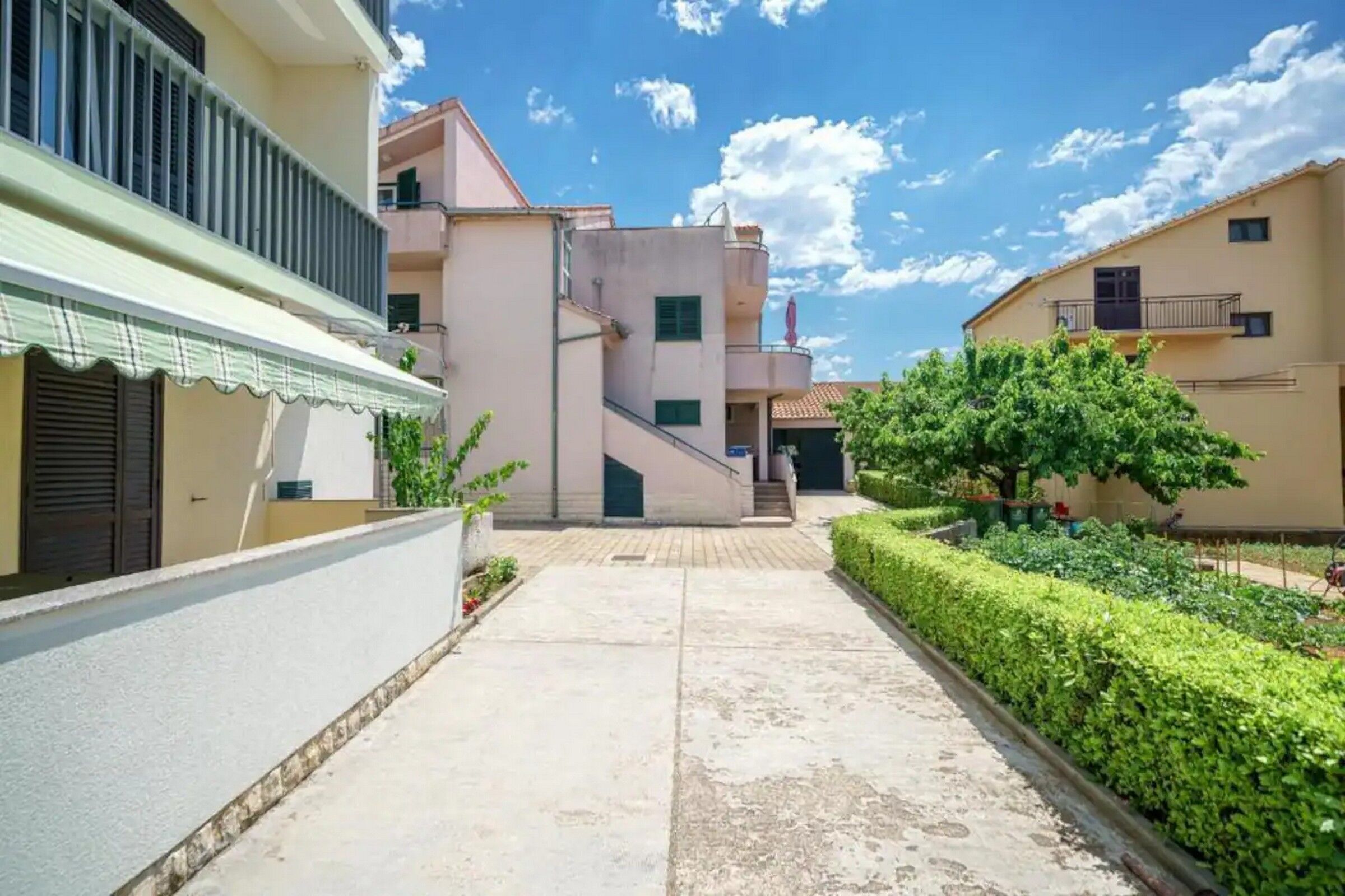 A residential area with a paved pathway, buildings with balconies, and a well-maintained hedge.