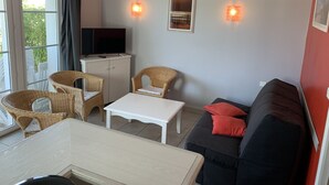 A living room with a white table, two wicker chairs, a black sofa with red cushions, a TV on a cabinet, and two wall-mounted lights. - Family home in the heart of Domaine Belle Dune (Fort-Mahon-Plage)