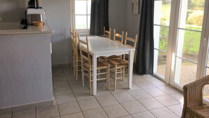 A kitchen with a coffee maker, a dining table with chairs, and a view of the outdoors through a glass door. - Family home in the heart of Domaine Belle Dune (Fort-Mahon-Plage)