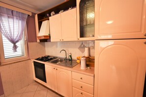 A kitchen with white cabinets, a stove, a sink, and a window with a sheer curtain. - Apartment Granum (Biograd na Moru)