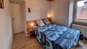 Une chambre avec un plafond en bois, un lit recouvert d’une couvre-lit à motifs, une table de chevet et une fenêtre donnant sur un toit recouvert de tuiles.