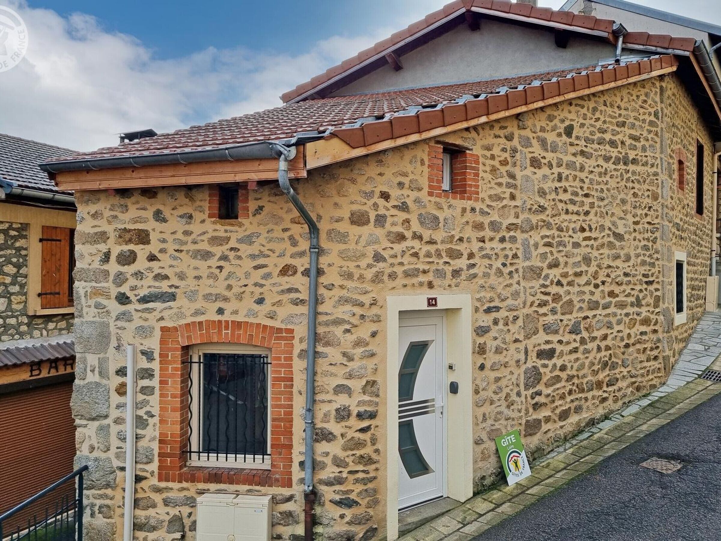 Un bâtiment en pierre avec un toit en tuiles, une porte blanche et une petite fenêtre ornée d’une grille métallique.