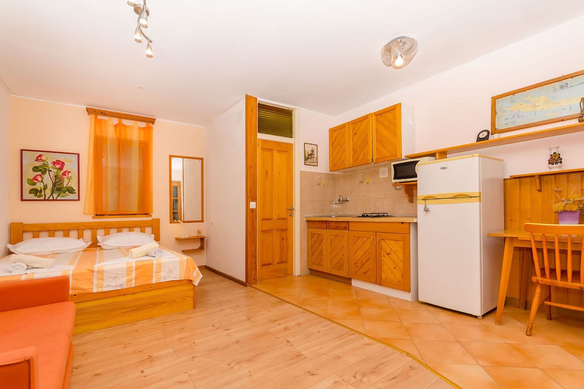 A room with a bed, a small table, a chair, a kitchen with wooden cabinets, a refrigerator, and a window with curtains.