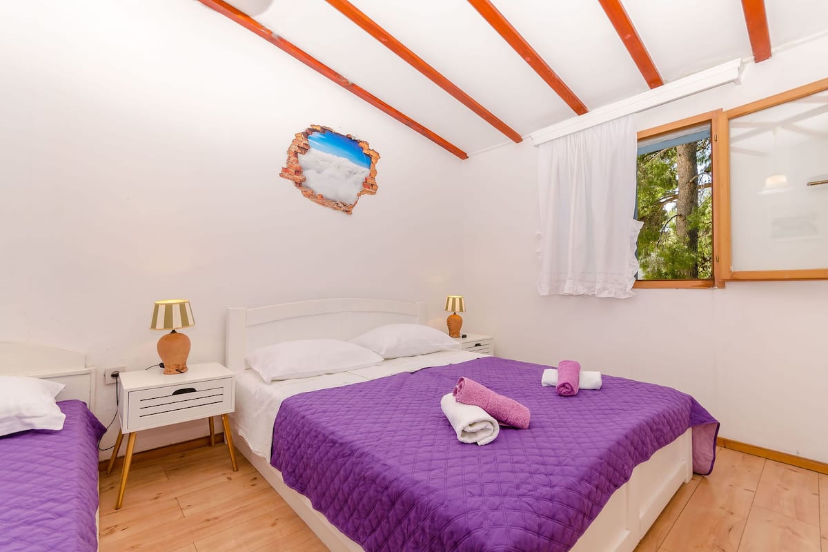 A bedroom with a large bed, two bedside tables, a window with a view of trees, and a ceiling with wooden beams.
