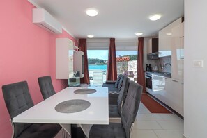 A modern kitchen with white cabinets, a dining area with a white table and chairs, and a view of the sea through large windows. - Apartments Villa Summertime (Tisno)