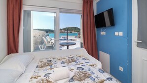 Habitación de hotel con cama, televisor montado en la pared y puerta de vidrio corredera que da a un balcón con vistas a la playa y las colinas.
