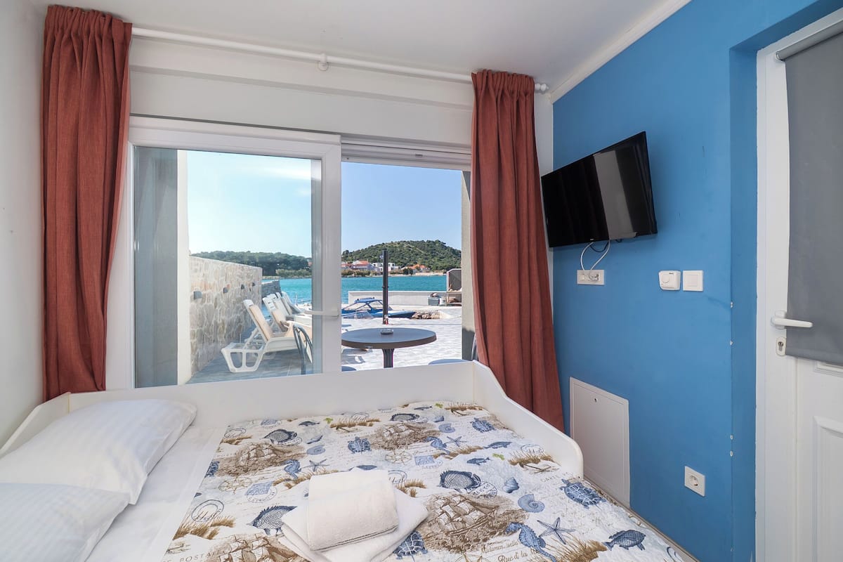 Ett hotellrum med en säng, en tv som är monterad på väggen och en skjutdörr i glas som leder ut till en balkong med utsikt över en strand och kullar.