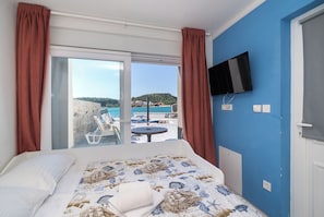 Ett hotellrum med en säng, en tv som är monterad på väggen och en skjutdörr i glas som leder ut till en balkong med utsikt över en strand och kullar.