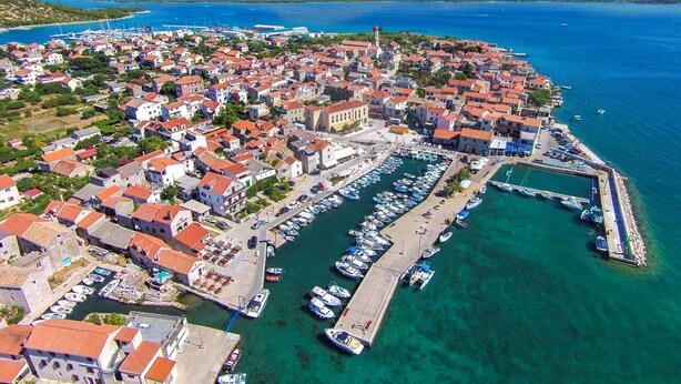 Vista aérea de un pueblo costero con un puerto deportivo, casas con tejados rojos y una iglesia destacada.