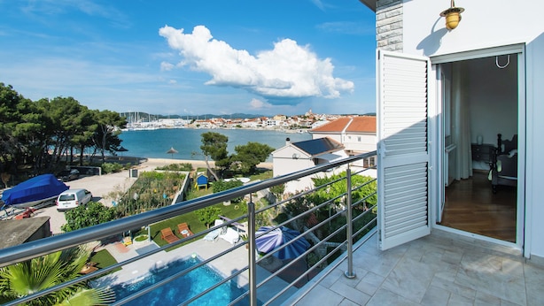 A balcony with a view of a beach and sea, a swimming pool, and a room with a bed and a window. - Apartment Villa Zlatna (Tisno)