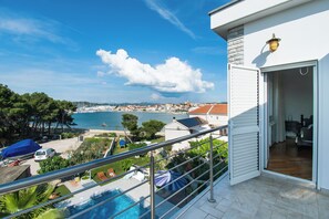 A balcony with a view of a beach and sea, a swimming pool, and a room with a bed and a window. - Apartment Villa Zlatna (Tisno)