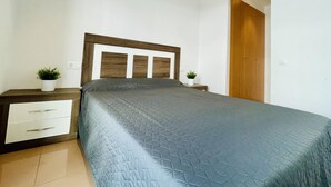 Un dormitorio con cama, cabecera de madera, mesitas de noche con plantas, un armario y suelo de baldosas.
