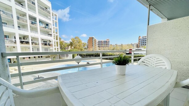 Un balcón con una mesa y sillas blancas, una planta en maceta y vistas a una piscina y edificios.