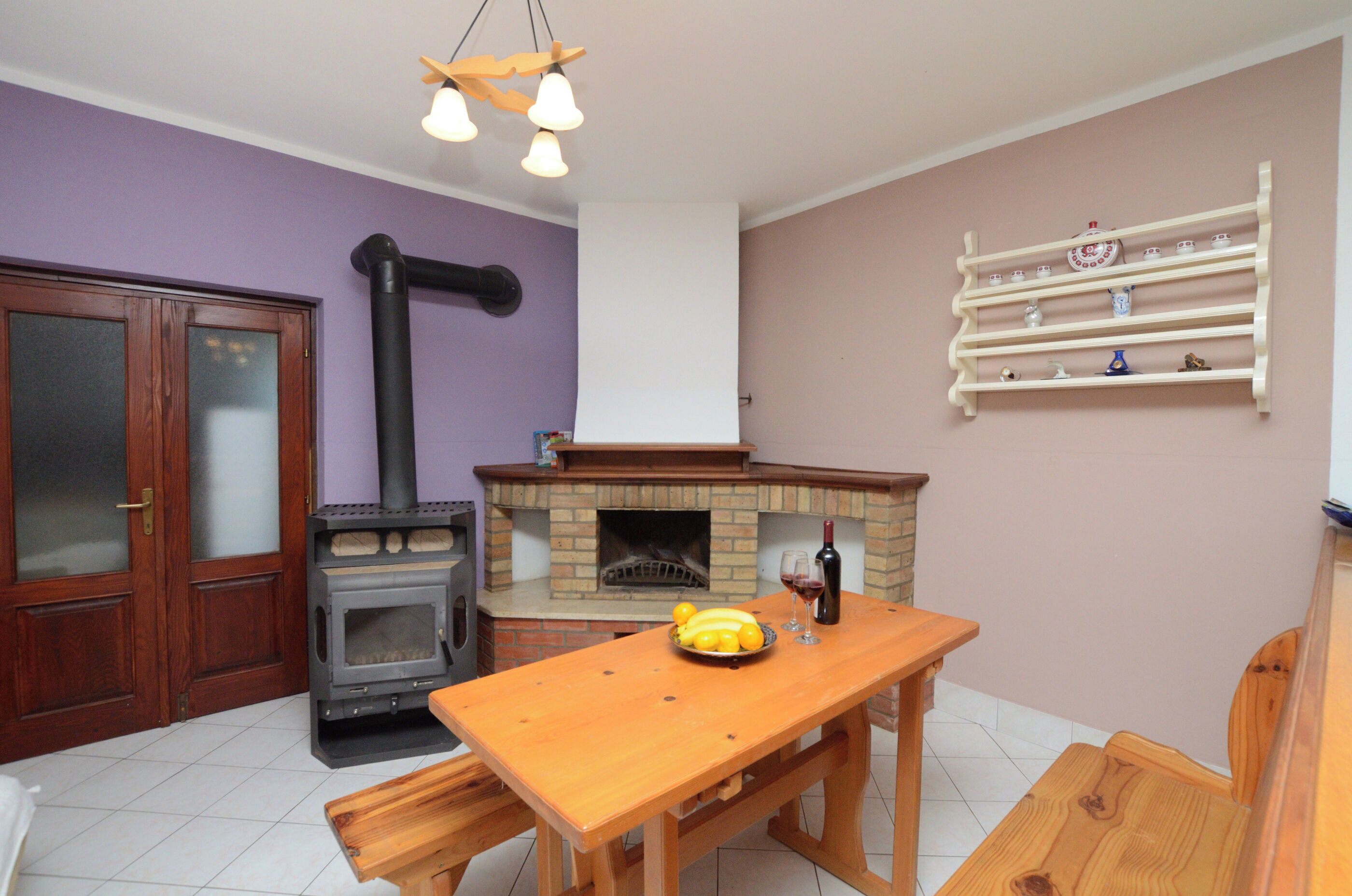 A kitchen with a wood-burning stove, a dining table, and a shelf with kitchenware.