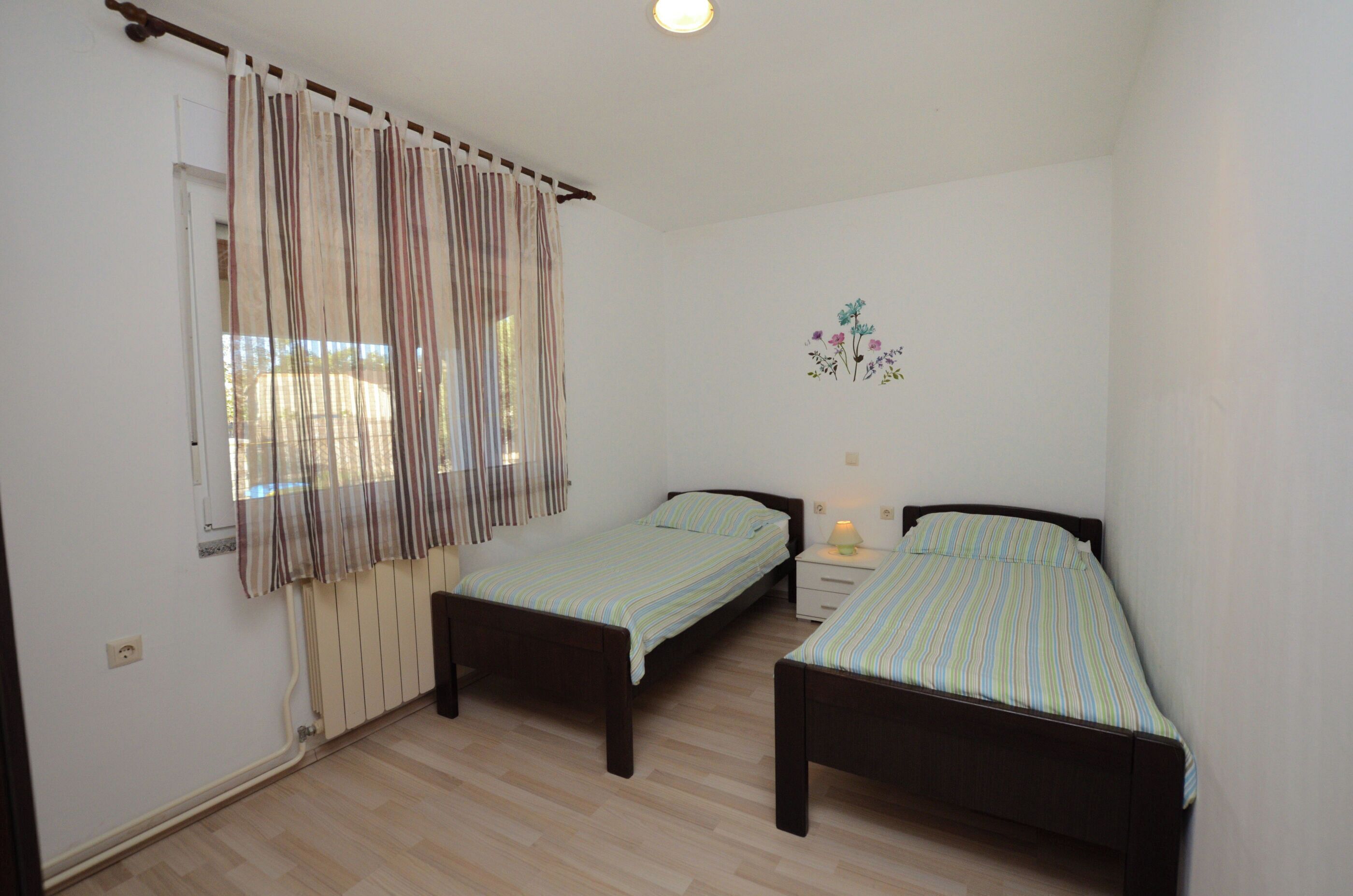 A small, clean room with two single beds, a window with curtains, a small bedside table with a lamp, and a wall decoration.