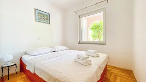 A bedroom with a bed, pillows, a window, a small table, and a framed picture on the wall. - Apartment Brdar (Crikvenica)