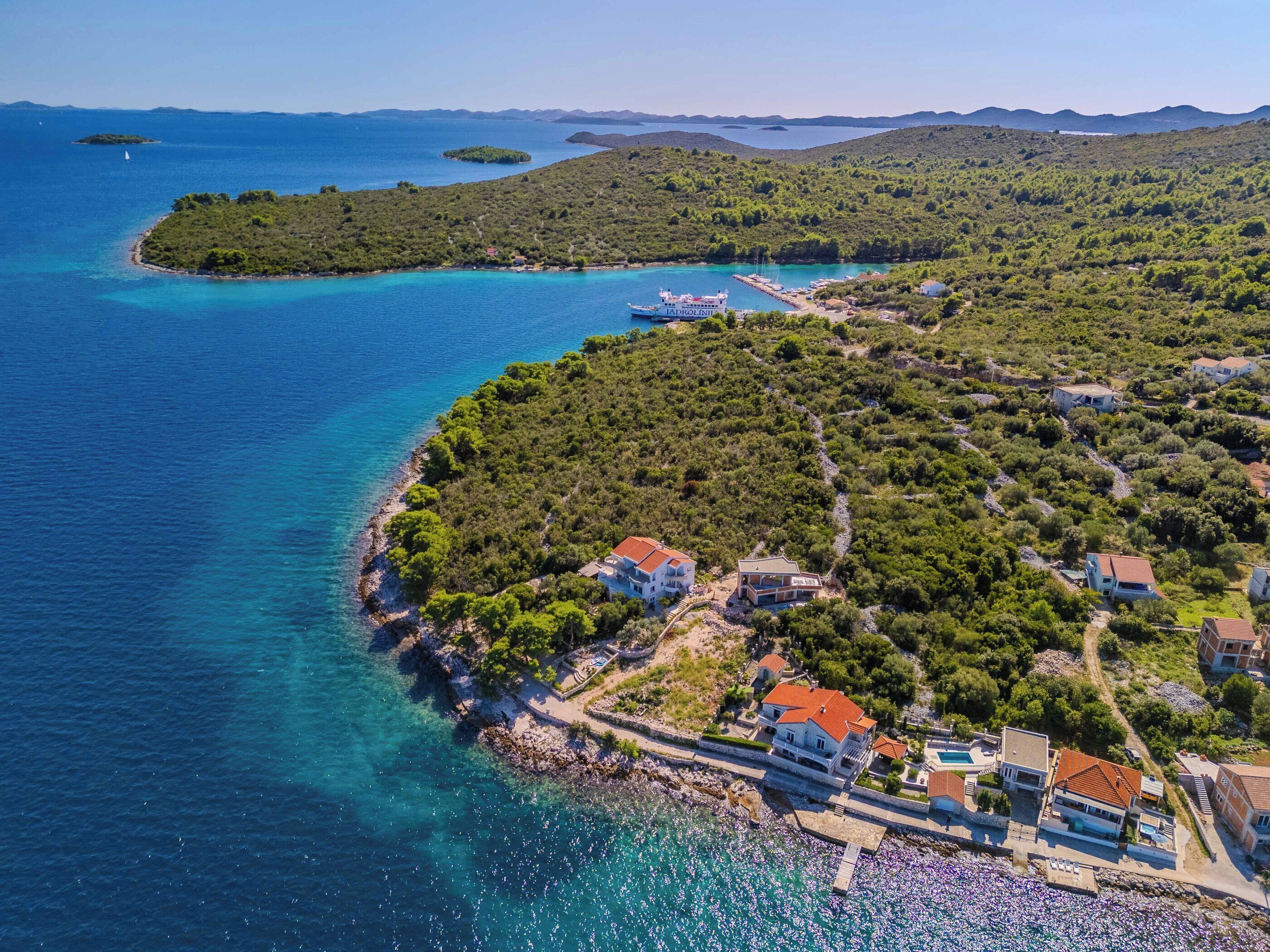 Ein Luftbild eines Küstendorfes mit Häusern entlang der Uferlinie, einem klaren blauen Meer und einer hügeligen Landschaft im Hintergrund.