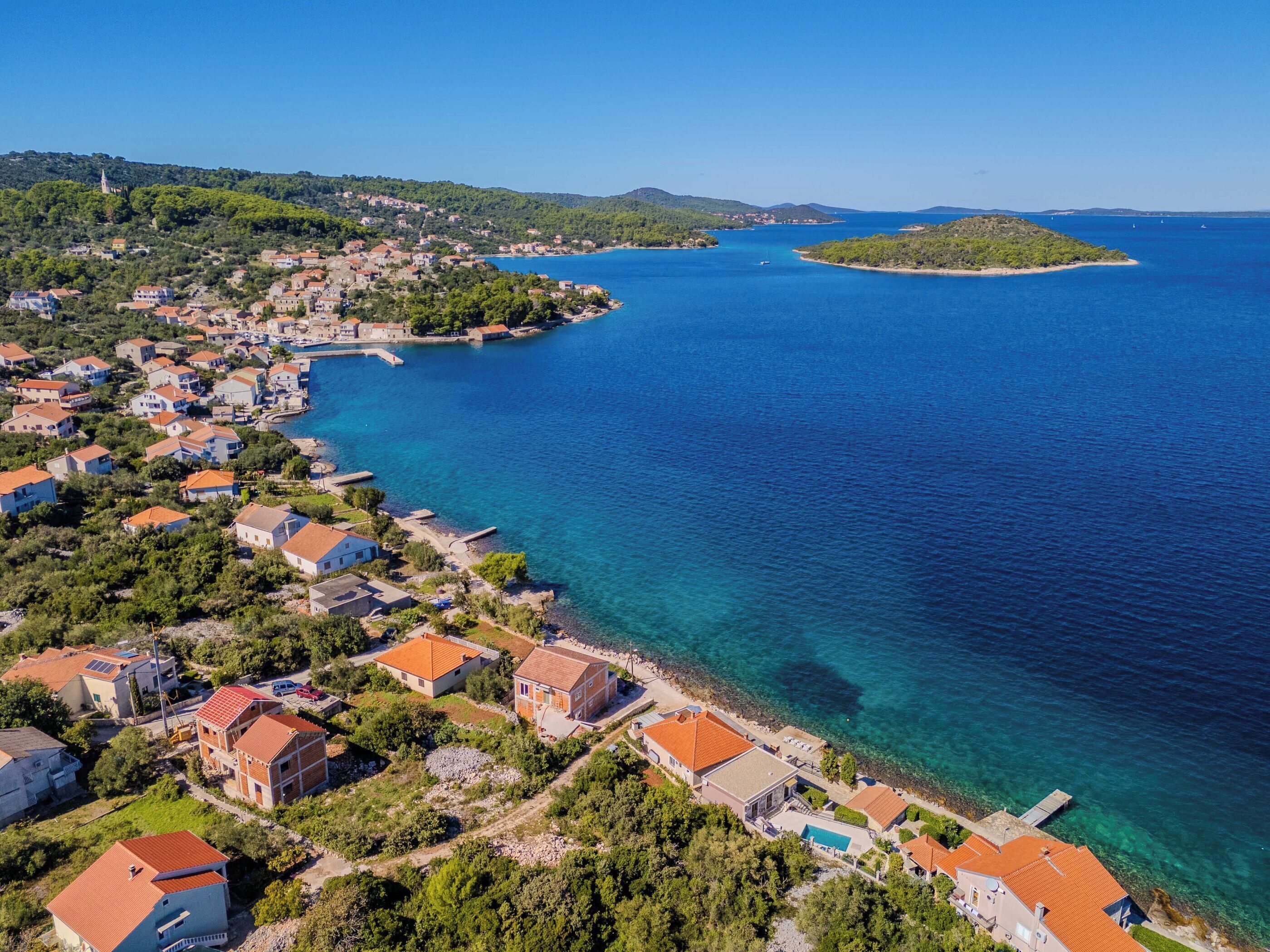 An aerial view of a coastal town with houses along the shoreline, a clear blue sea, and a small island in the distance.