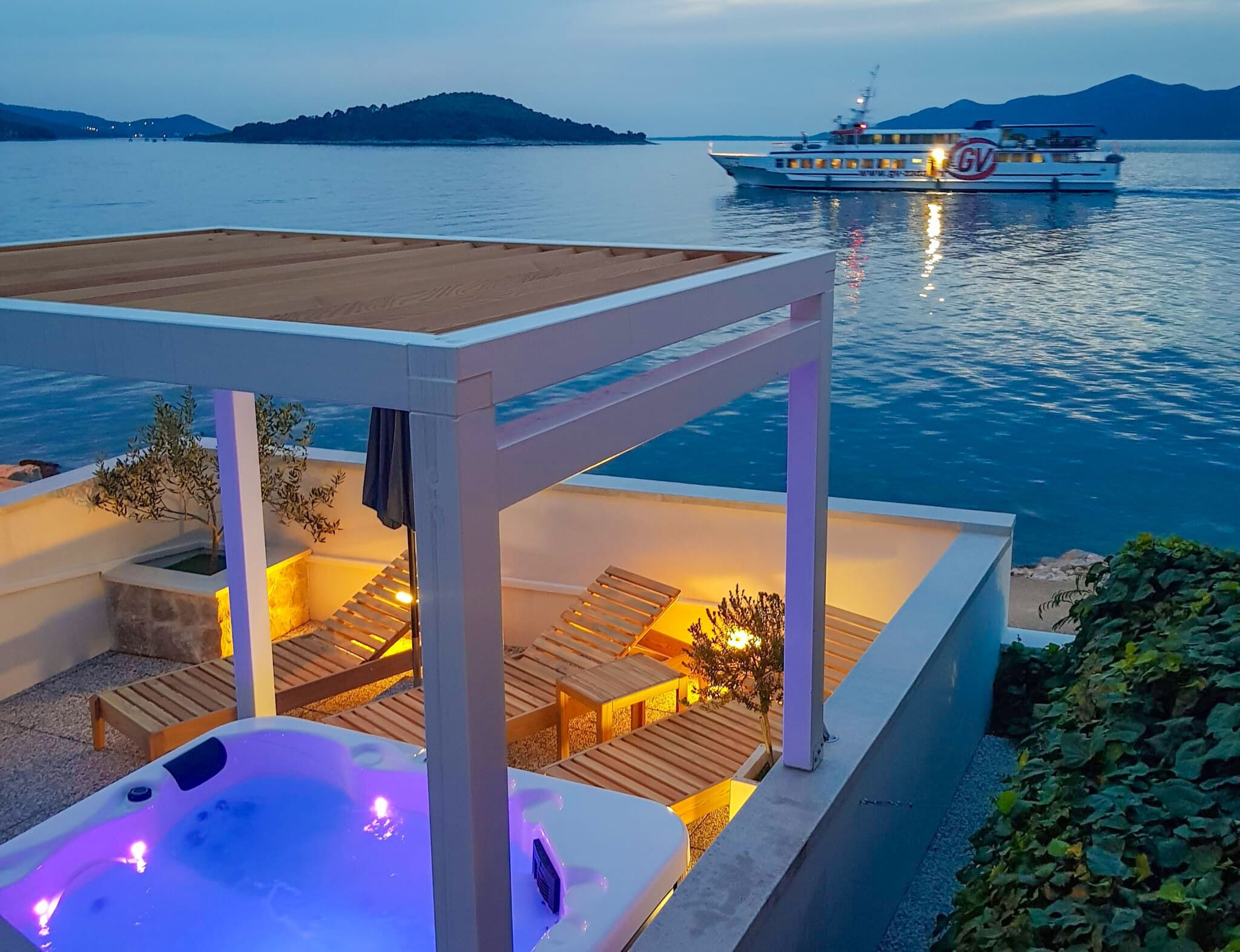 A hot tub with a wooden deck and lounge chairs, a boat with 'GV' on its side, and a mountainous landscape in the background.