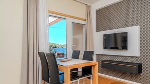 Ein moderner Essbereich mit Holztisch und schwarzen Stühlen, einem an der Wand montierten Fernseher und Meerblick durch ein großes Fenster.