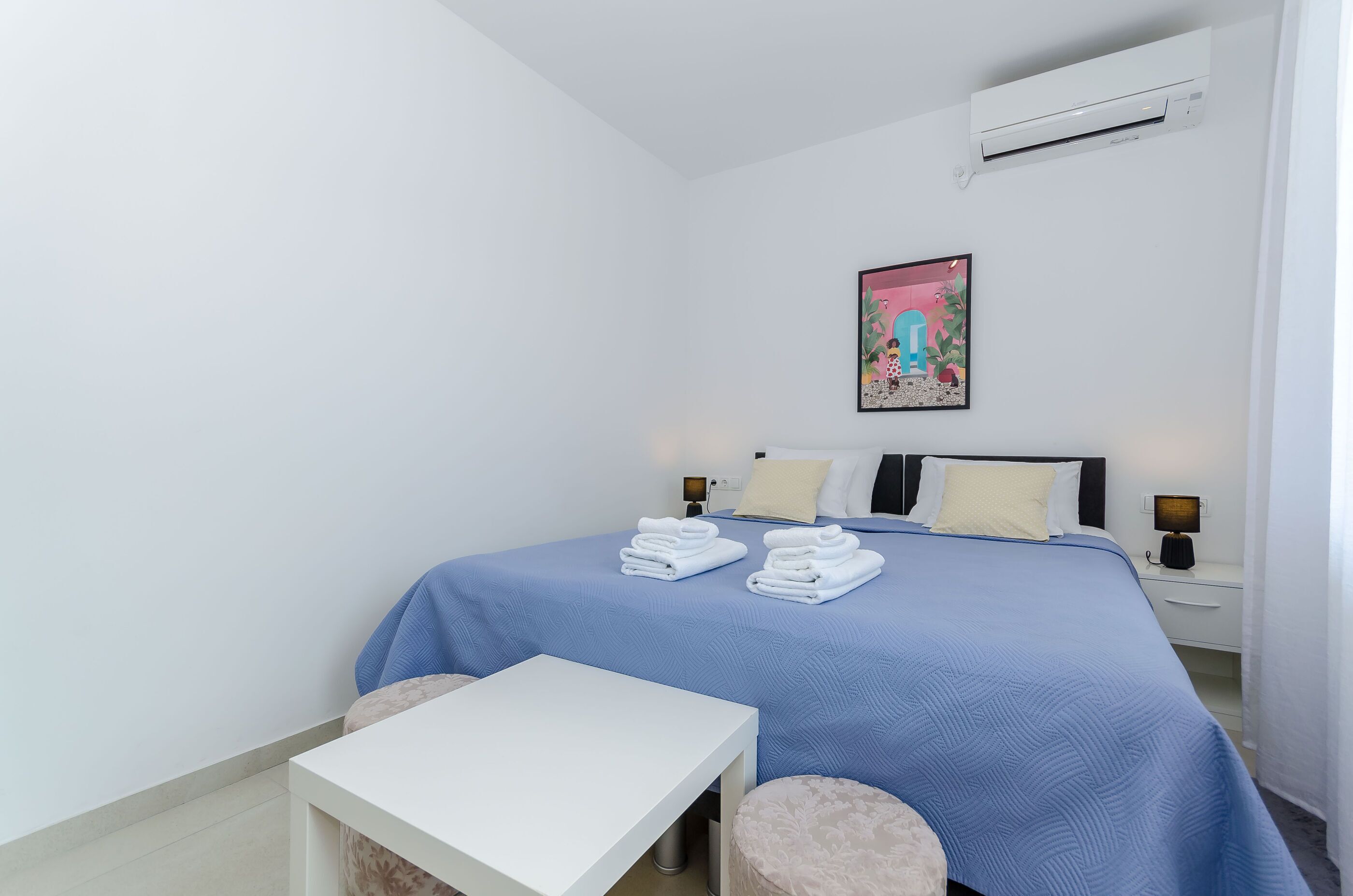 A bedroom with a bed, bedside tables, a table, and an air conditioning unit.