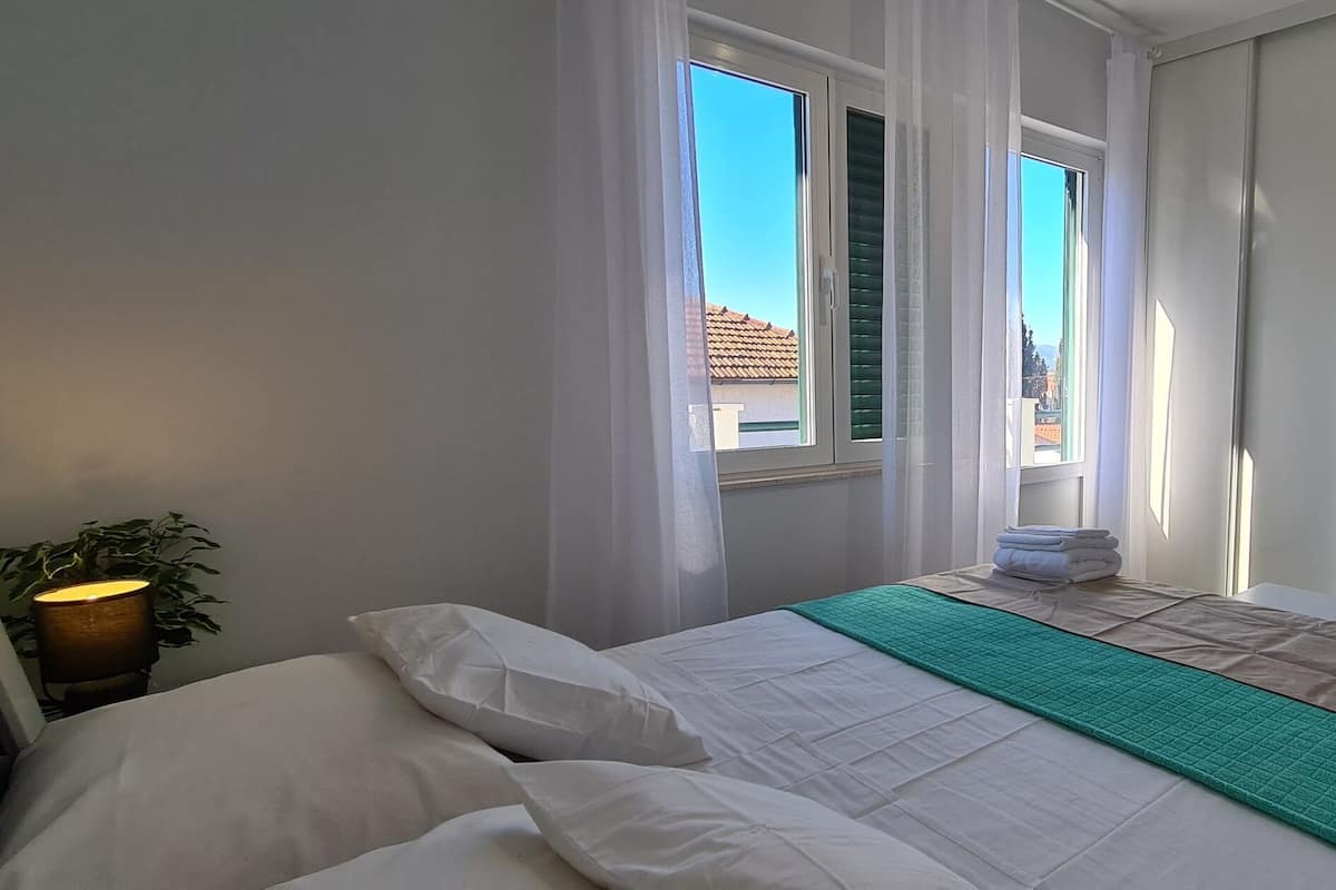 Une chambre à coucher avec un lit, une table de chevet avec une lampe, un téléviseur fixé au mur et une fenêtre avec des rideaux.