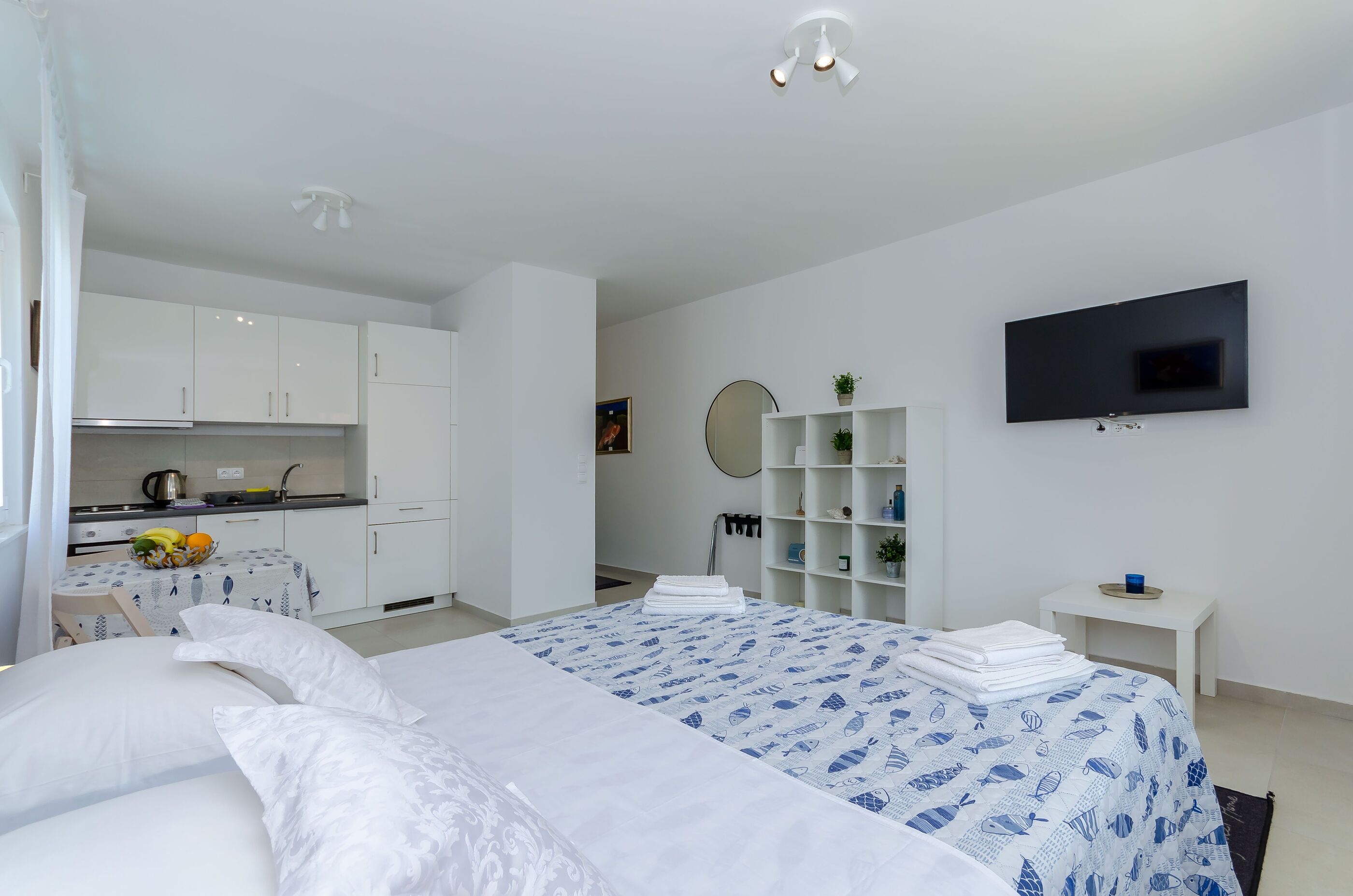A modern hotel room with a large bed, a TV mounted on the wall, a small kitchen area, and a bedside table with a plant.