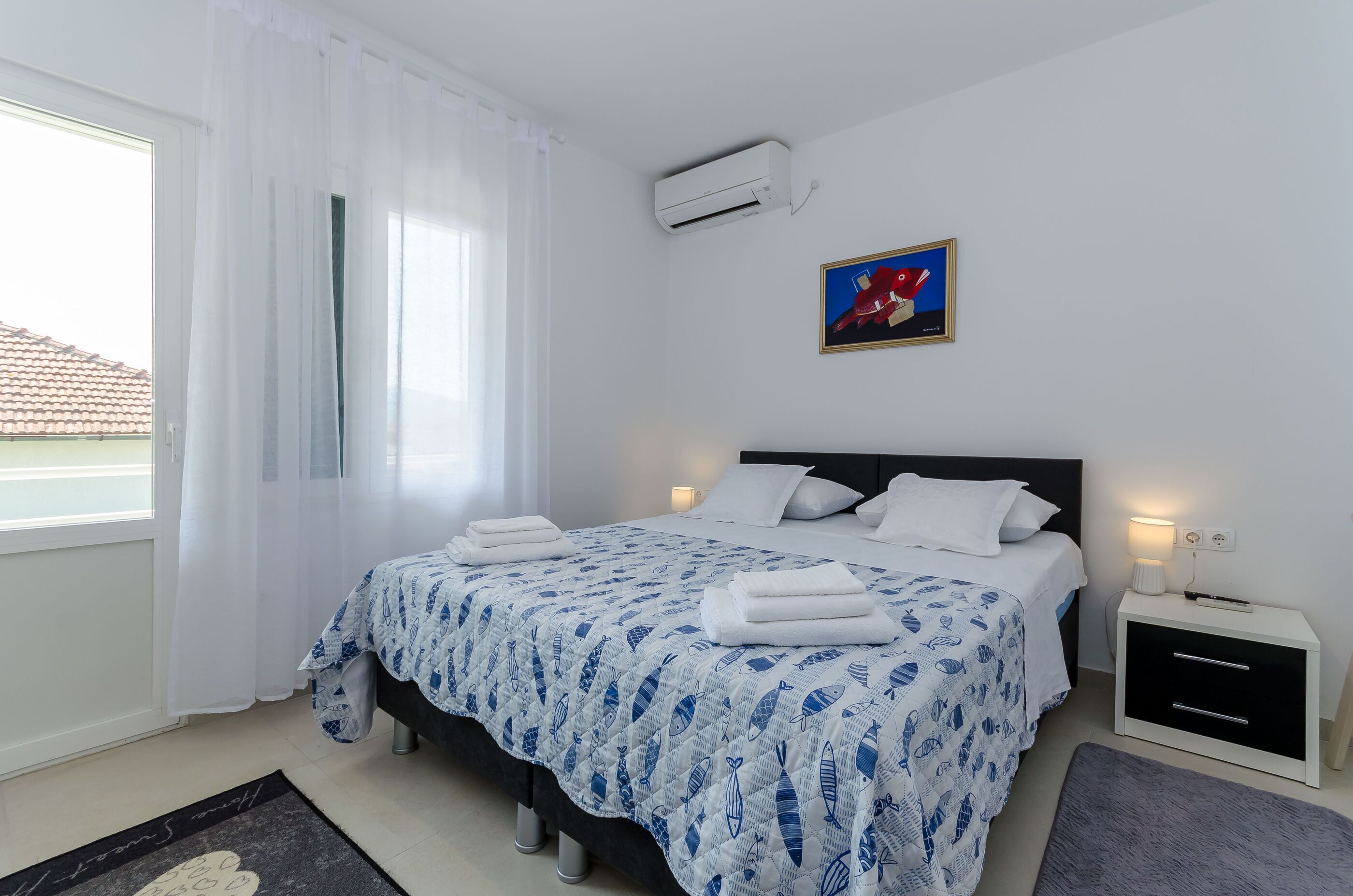 A bedroom with a bed, white curtains, a painting, an air conditioner, a bedside table with a lamp, and a small rug.