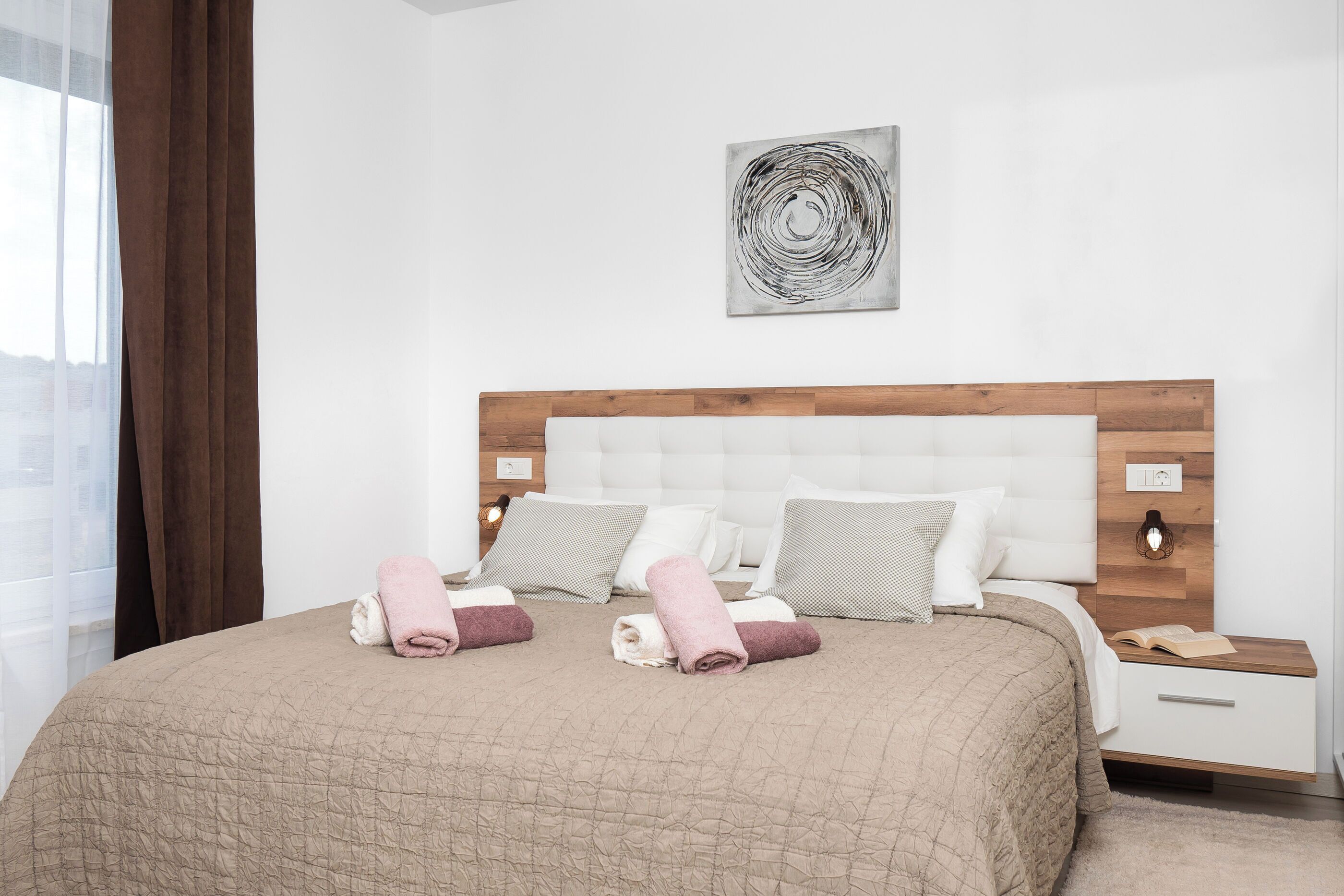 A neatly made bed with a beige quilt, pillows, and rolled towels. There is a wooden headboard, a bedside table with a lamp, and a wall art piece above the bed.