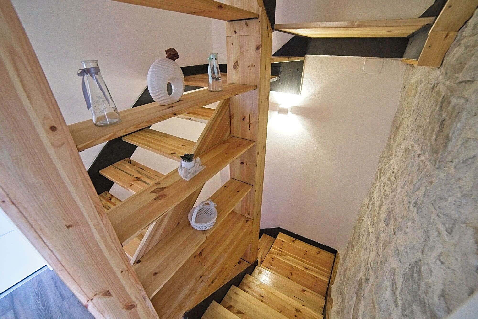 A wooden staircase with a rustic design, leading to a room with a stone wall and a small shelf with decorative items.