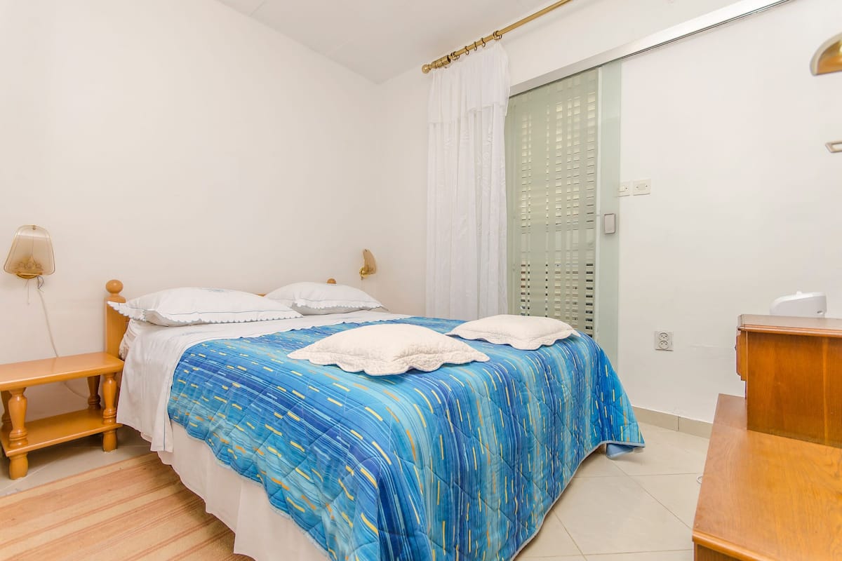 Ein Schlafzimmer mit einem Bett, Nachttischen, einer hölzernen Kommode, einem Fenster mit Vorhängen und einer Wandleuchte.