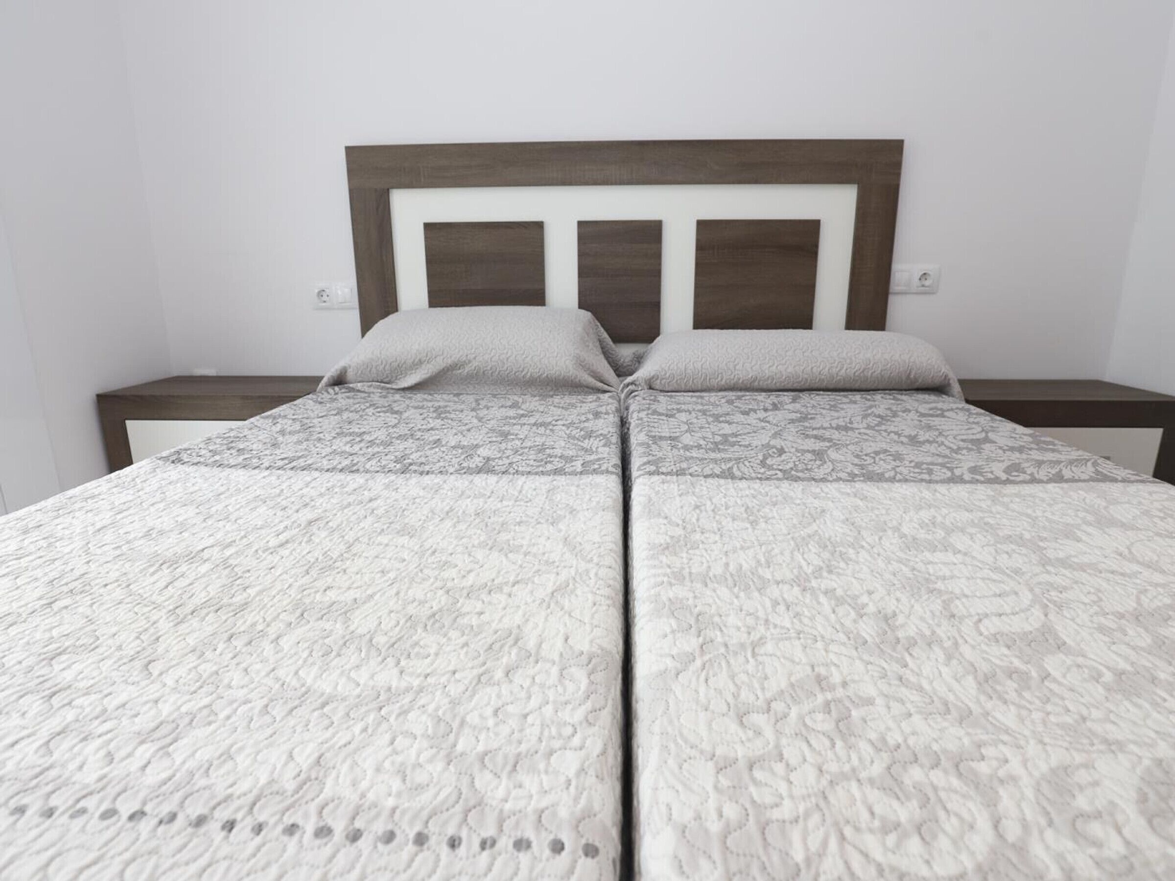Uma cama de casal com cabeceira de madeira, duas mesas de cabeceira e um cobreleite estampado.