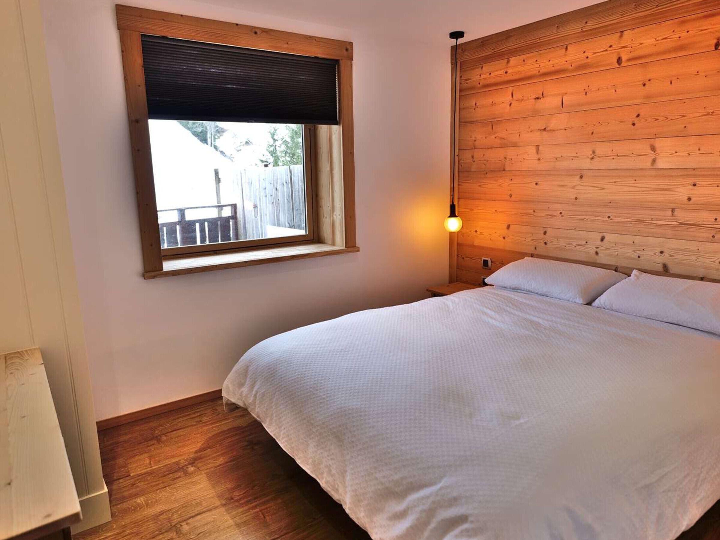 Quarto com cabeceira de madeira, janela com persiana, abajur e cama com roupa de cama branca.