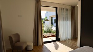 Una habitación de hotel moderna con un amplio ventanal corredero que da a un balcón, una silla cómoda y un pequeño reposapiés.